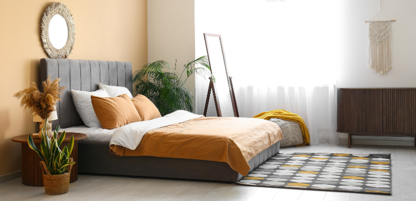 A modern bedroom with a gray bed, mustard yellow bedding, green plants, and warm beige walls, accented by a mirror and macrame decor.