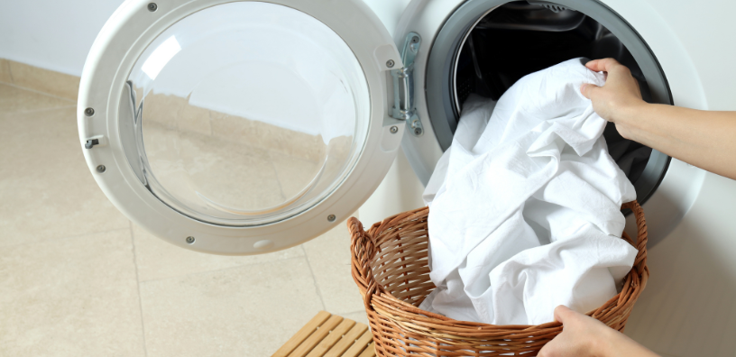 A person is placing a freshly laundered white sheet into a front-loading washing machine.