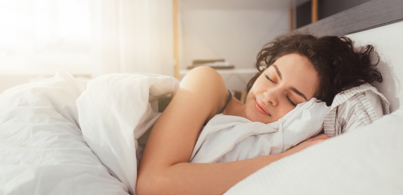 Young woman peacefully sleeping in bed, wrapped in cozy white bedding with a soft smile on her face, bathed in gentle morning light filtering through a nearby window.