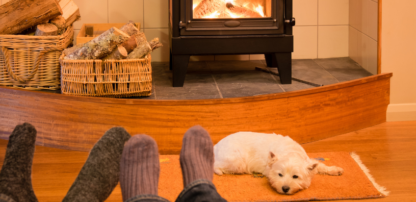 A tranquil and heartwarming scene of a small white dog resting on a cozy rug in front of a roaring fireplace, with two people’s feet in warm socks enjoying the warmth.