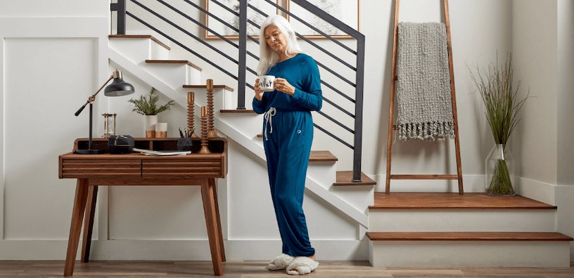 A woman in a blue loungewear set relaxes at home. She is holding a white cup of coffee and standing near a set of stairs, likely in her living room or entryway.