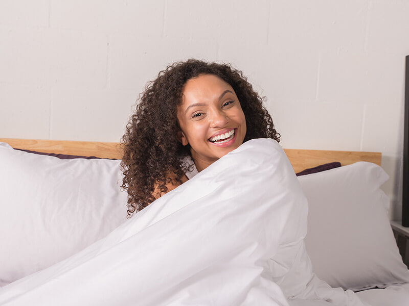 A woman smiles happily in bed, radiating warmth and comfort.