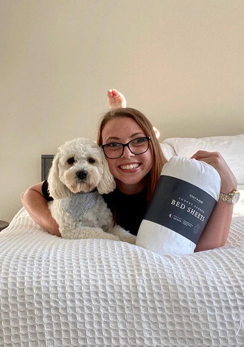 A smiling customer cuddling her dog and a packaged set of Cosy House Collection’s Luxury bed sheets on a cozy bed.